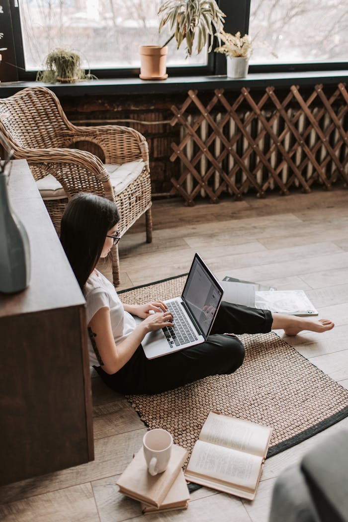 A young woman in casual attire working from home using a laptop while sitting on the floor with a coffee mug nearby.